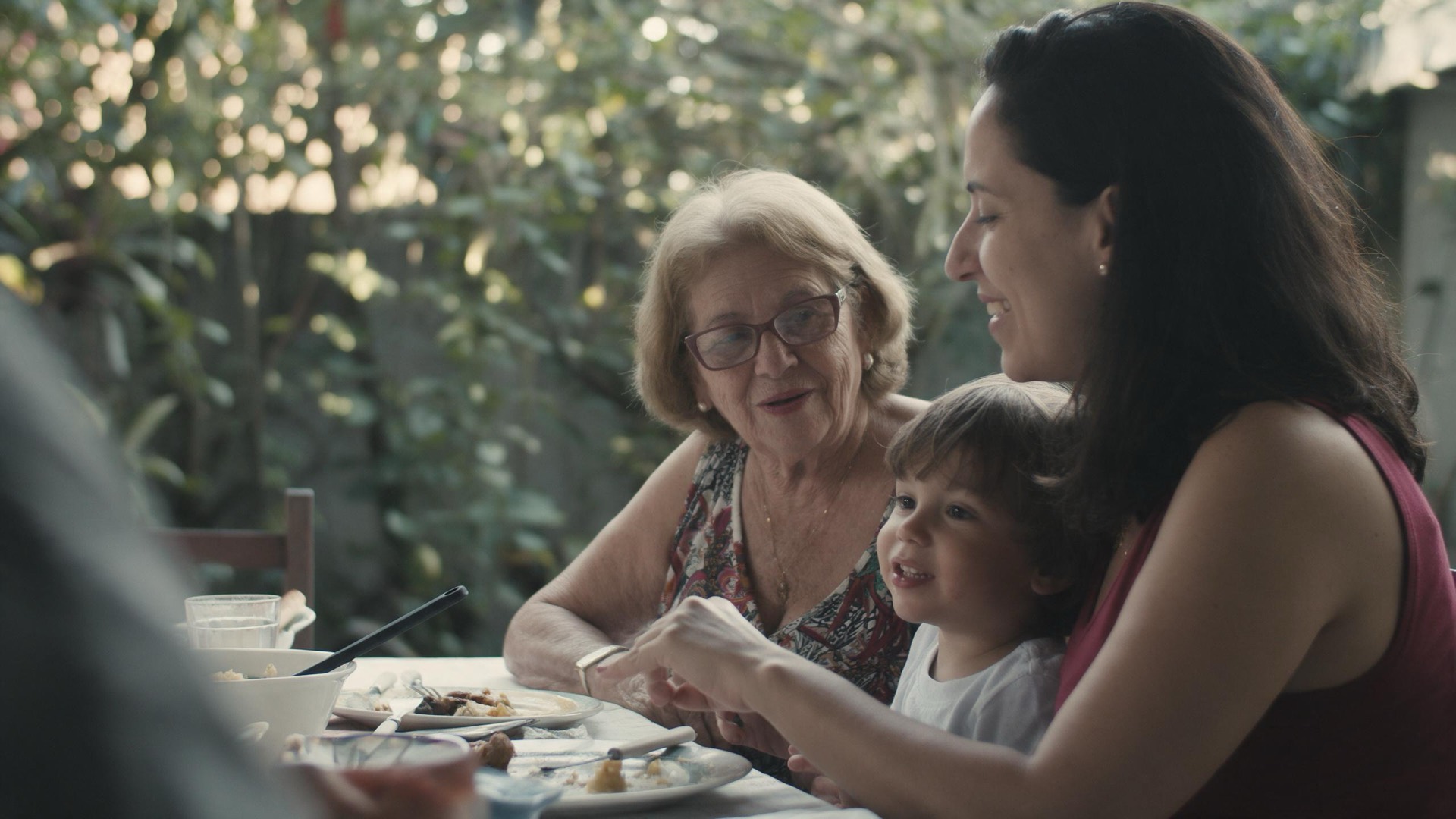 Família de três gerações reunida à mesa em um quintal arborizado